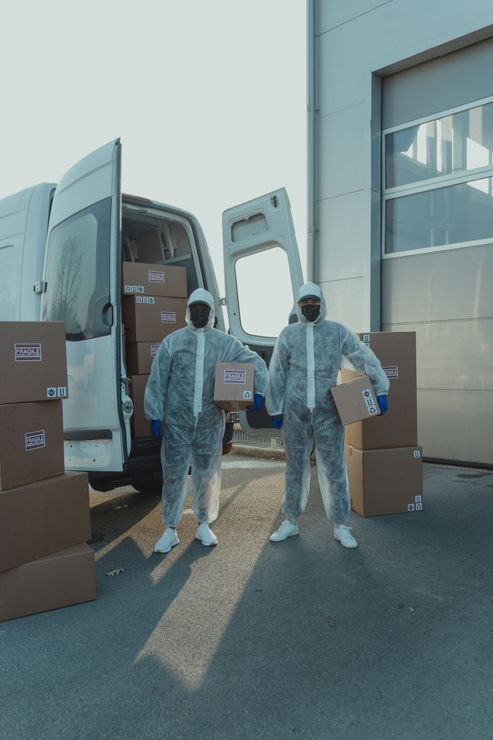 Two workers in protective suits load cardboard boxes into a van outdoors.